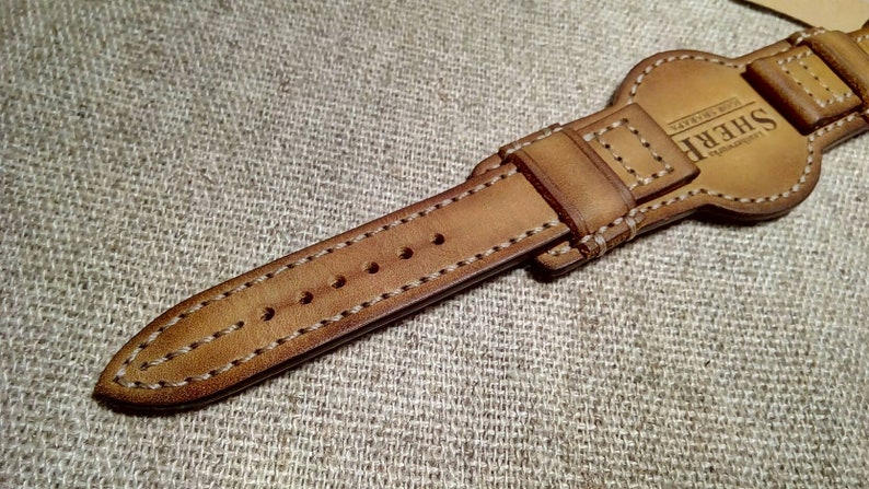 May include: A close-up shot of a tan leather watch strap with white stitching. The strap features a buckle and adjustment holes. The brand name "SHERE" is visible. The background is a textured, neutral-toned fabric.