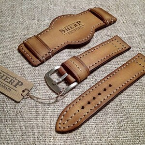 May include: A collection of light brown leather watch straps with white stitching. The set includes a strap, a buckle, and a watch back piece. A wooden tag with the text "SHERP Leatherworks IGOR SHARAPA" is also included.
