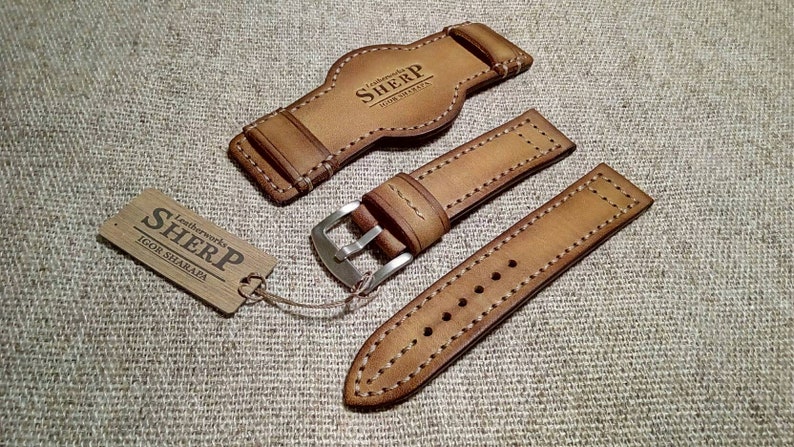 May include: A collection of light brown leather watch straps with white stitching. The set includes a strap, a buckle, and a watch back piece. A wooden tag with the text "SHERP Leatherworks IGOR SHARAPA" is also included.