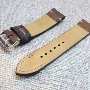 May include: A brown leather watch strap with white stitching and a silver buckle. The strap is shown on a beige fabric background.