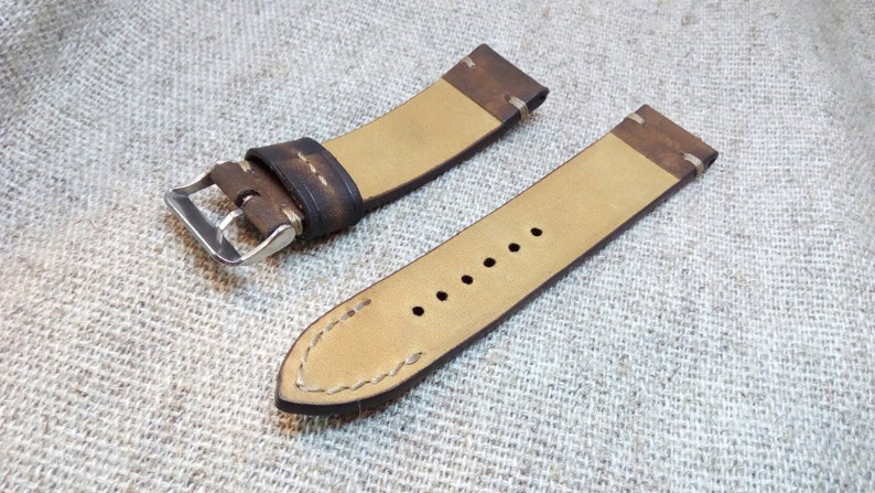May include: A brown leather watch strap with white stitching and a silver buckle. The strap is shown on a beige fabric background.