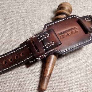 May include: A brown leather watch strap with white stitching and a silver buckle. The strap has a leather keeper with the text "Handmade" embossed on it.