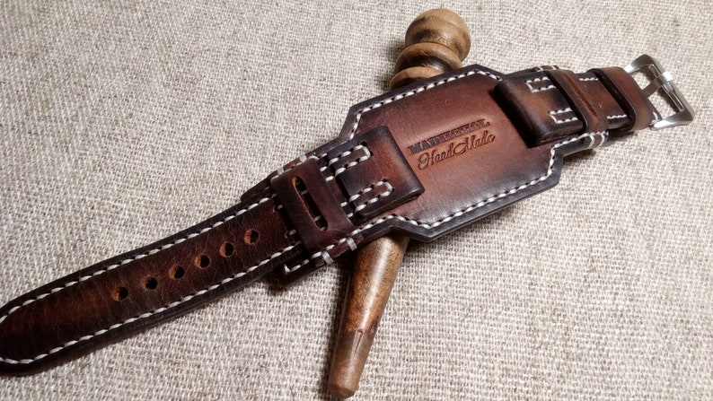May include: A brown leather watch strap with white stitching and a silver buckle. The strap has a leather keeper with the text "Handmade" embossed on it.