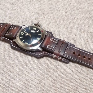 May include: A brown leather watch strap with white stitching. The strap is attached to a silver watch with a black face and white numbers.