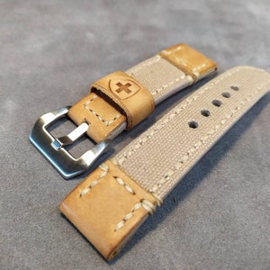 May include: A beige canvas watch strap with brown leather accents and a silver buckle. The strap has a stitched design and a small cross logo on the leather.