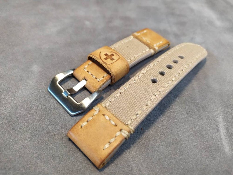 May include: A beige canvas watch strap with brown leather accents and a silver buckle. The strap has a stitched design and a small cross logo on the leather.