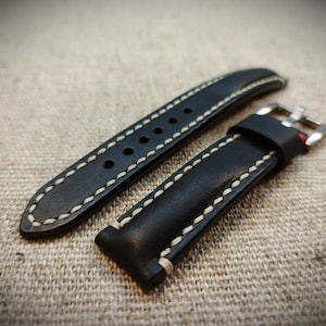 Könnte beinhalten: Zweiteiliges schwarzes Lederarmband mit cremefarbenen Nähten. Das Armband hat eine silberfarbene Schnalle und mehrere Löcher zur Anpassung. Das Leder scheint hochwertig und langlebig zu sein, passend für eine klassische Uhr.