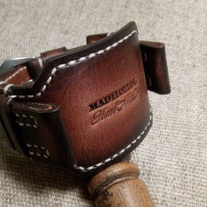 May include: A brown leather watch band with white stitching. The band has a buckle and is labeled "Madrerool Hand Made".
