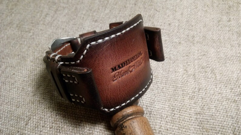 May include: A brown leather watch band with white stitching. The band has a buckle and is labeled "Madrerool Hand Made".