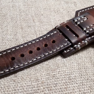 May include: A brown leather watch strap with white stitching and multiple holes for adjusting the size. The strap has a buckle with the text "Hand Made" on it.