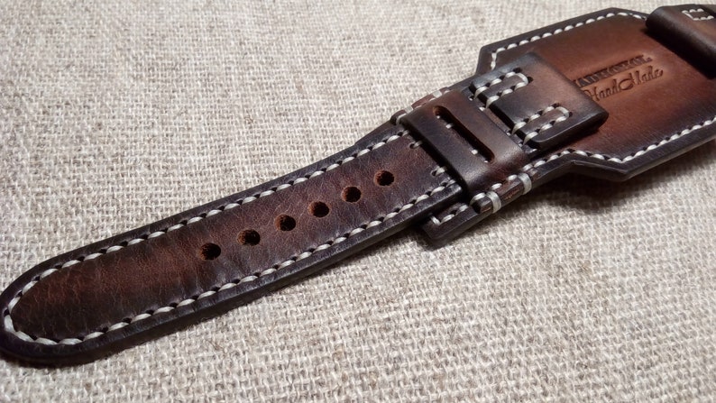 May include: A brown leather watch strap with white stitching and multiple holes for adjusting the size. The strap has a buckle with the text "Hand Made" on it.