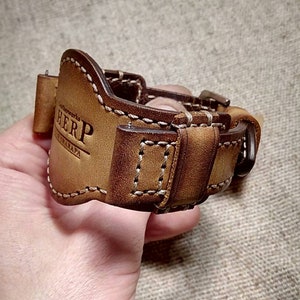May include: A brown leather watch strap with white stitching and a buckle. The strap has the text "HERP" and "CATALAPA" embossed on it. The watch strap is designed to fit a watch. The leather has a distressed appearance.