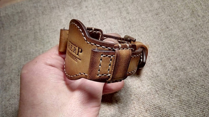 May include: A brown leather watch strap with white stitching and a buckle. The strap has the text "HERP" and "CATALAPA" embossed on it. The watch strap is designed to fit a watch. The leather has a distressed appearance.