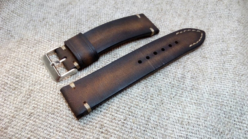 May include: A brown leather watch strap with white stitching. The strap has a silver buckle and is designed for a wristwatch.
