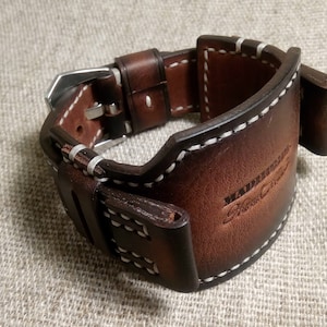 May include: A brown leather watch strap with white stitching and a silver buckle. The strap has the text "Handmade Fine Crafts" embossed on it.
