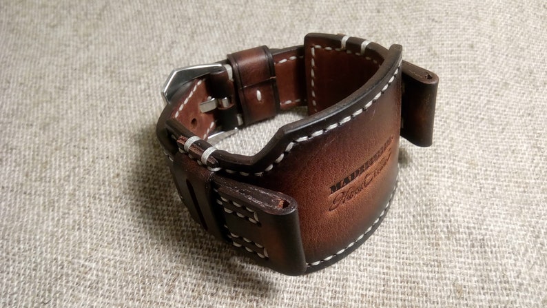 May include: A brown leather watch strap with white stitching and a silver buckle. The strap has the text "Handmade Fine Crafts" embossed on it.