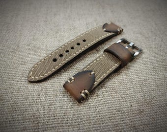 May include: A two-piece watch strap featuring a tan canvas center section with light-colored stitching. The ends are made of dark brown leather with a distressed finish and matching stitching. A silver-toned metal buckle is attached to one end.