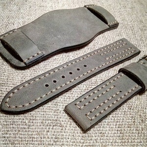 May include: A grey leather watch strap set consisting of a wide bund pad, a long strap with multiple adjustment holes, and a shorter strap with a metal buckle. The pieces feature visible tan stitching along the edges and a small branded tag.