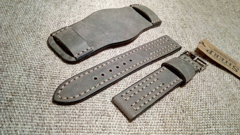 May include: A grey leather watch strap set consisting of a wide bund pad, a long strap with multiple adjustment holes, and a shorter strap with a metal buckle. The pieces feature visible tan stitching along the edges and a small branded tag.