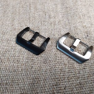 May include: Two watch band buckles, one black and one silver, on a textured fabric background.