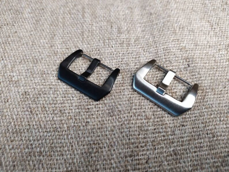 May include: Two watch band buckles, one black and one silver, on a textured fabric background.