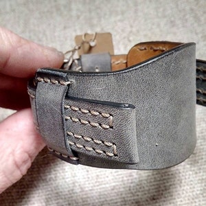 May include: A wide gray leather cuff bracelet featuring a smooth matte finish, dark edges, and visible light-colored stitching along the seams. The bracelet includes a matching leather loop and a small brown paper tag attached to the side.