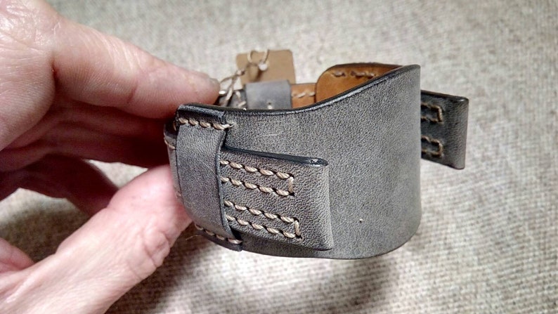 May include: A wide gray leather cuff bracelet featuring a smooth matte finish, dark edges, and visible light-colored stitching along the seams. The bracelet includes a matching leather loop and a small brown paper tag attached to the side.