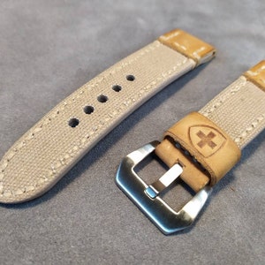 May include: Two beige canvas watch straps with brown leather accents. One strap has a silver buckle and the other has a small cross logo on the leather.