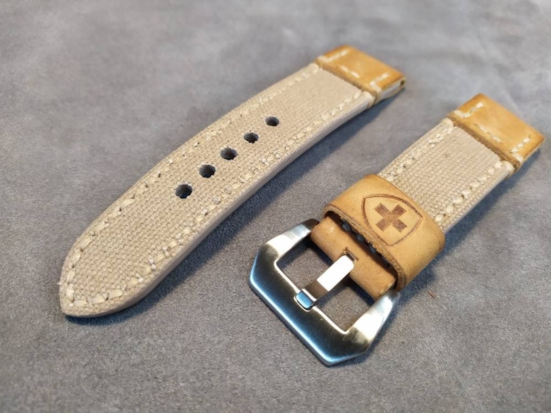 May include: Two beige canvas watch straps with brown leather accents. One strap has a silver buckle and the other has a small cross logo on the leather.
