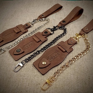 Pode incluir: Porta-chaves de couro castanho com detalhes em corrente de metal. Cada porta-chaves apresenta uma tira de couro com fecho de pressão e a palavra "SHERP" em relevo. Correntes prateadas, pretas e douradas.