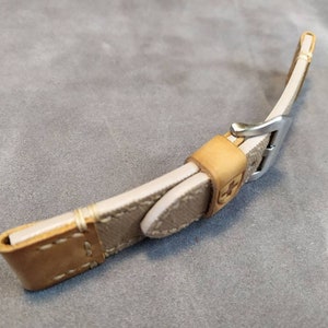May include: A beige leather watch strap with a silver buckle. The strap has a stitched design and a brown leather lining.