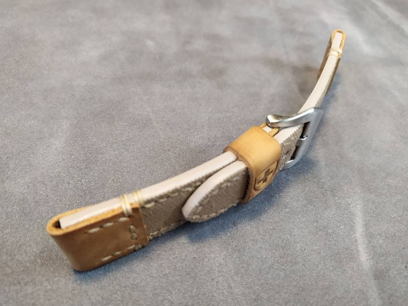 May include: A beige leather watch strap with a silver buckle. The strap has a stitched design and a brown leather lining.