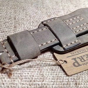 May include: A grey leather watch strap featuring a wide cuff design with light-colored contrast stitching, a silver-toned metal buckle, and a rectangular wooden tag attached by a string that reads Leatherworks SHERP IGOR SHARAPA.