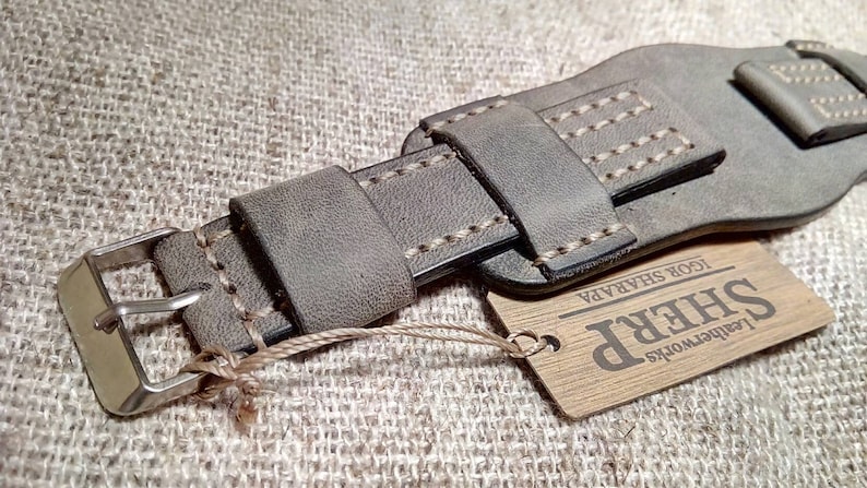 May include: A grey leather watch strap featuring a wide cuff design with light-colored contrast stitching, a silver-toned metal buckle, and a rectangular wooden tag attached by a string that reads Leatherworks SHERP IGOR SHARAPA.