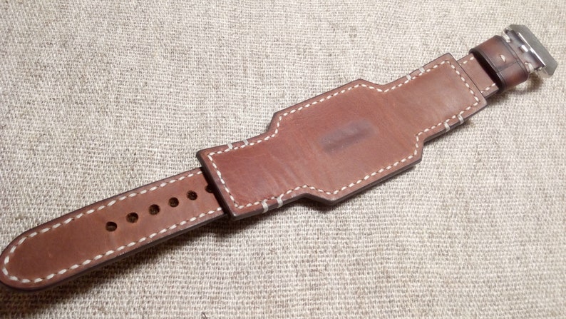 May include: Brown leather watch band with white stitching. The band has a buckle closure and is designed to fit a wrist.