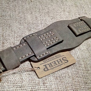 May include: A dark olive green leather watch strap featuring a wide cuff design with reinforced stitching and a silver-toned metal buckle. A rectangular wooden tag is attached by a string, displaying the text SHERP Leatherworks IGOR SHARAPA.