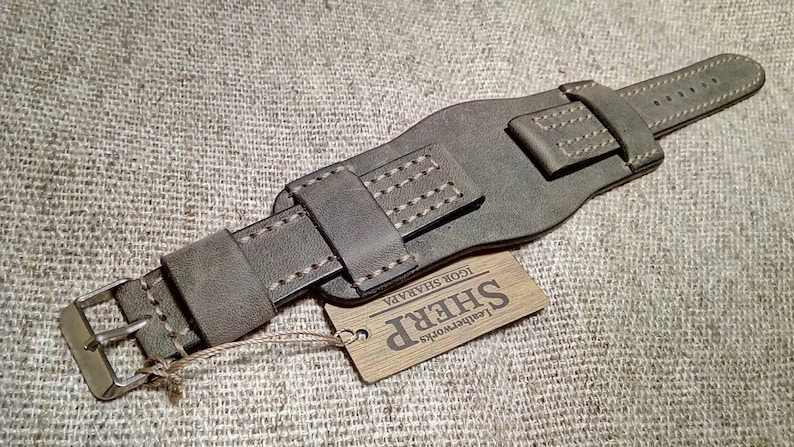 May include: A dark olive green leather watch strap featuring a wide cuff design with reinforced stitching and a silver-toned metal buckle. A rectangular wooden tag is attached by a string, displaying the text SHERP Leatherworks IGOR SHARAPA.