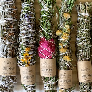 Op de afbeelding: Zes bundels gedroogde kruiden en bloemen die met touw aan elkaar zijn gebonden. De bundels zijn gelabeld met de tekst "timotea aromas de aras SAHUMERIO" en verschillende zinnen in het Spaans. De bundels zijn in een rij op een gestreept oppervlak gerangschikt.