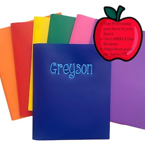May include: A stack of colorful folders, including orange, red, yellow, green, pink, blue, and purple. The front blue folder is embroidered with the name "Greyson" in a light blue script. An apple-shaped graphic with text is in the upper right corner.