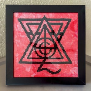 May include: A framed cross-stitch with a black geometric design on a red background.
