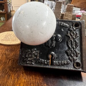May include: A white spherical glass doorknob attached to a rectangular black cast iron backplate. The metal plate features an ornate embossed pattern, a central keyhole, and the text JULY 21 1863 embossed near the bottom edge of the hardware.