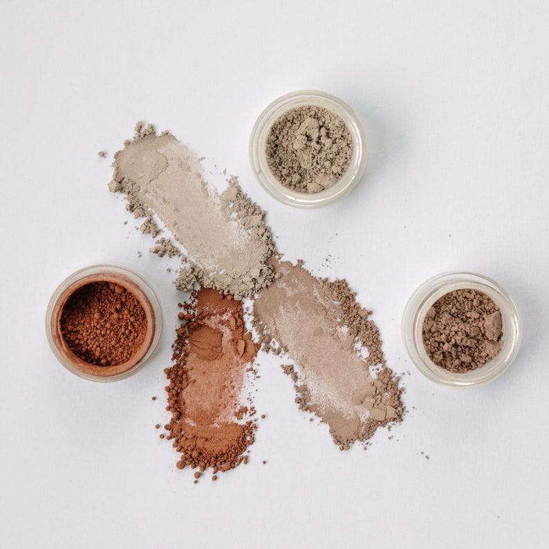 May include: Four small round containers of loose powder makeup in shades of brown, beige, and light brown. The powder is spilled out of the containers, creating streaks of color on a white background.