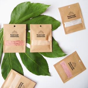 May include: Five small brown paper pouches with a sun and the text "honeypie minerals" on each. The pouches are arranged on a green leaf background.