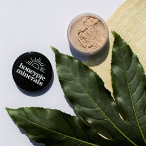May include: A black container with the text "honeypie minerals" and a sun graphic. The container is open and shows a light beige powder inside.
