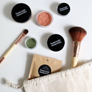 May include: A set of makeup products in a white canvas bag. The products include a green eyeshadow, a peach blush, a foundation, and a brush. The products are all labeled with the text "honeypie minerals".