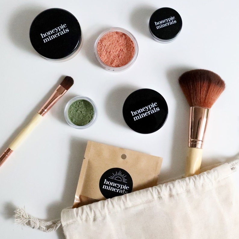 May include: A set of makeup products in a white canvas bag. The products include a green eyeshadow, a peach blush, a foundation, and a brush. The products are all labeled with the text "honeypie minerals".
