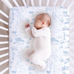 May include: A white crib with a white fitted sheet featuring a blue and white pattern of woodland animals and trees. A baby is sleeping on the sheet.