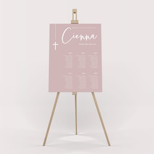 May include: A pink seating chart with a white cross and the name "Cienna" in cursive. The chart lists tables and names for a christening.