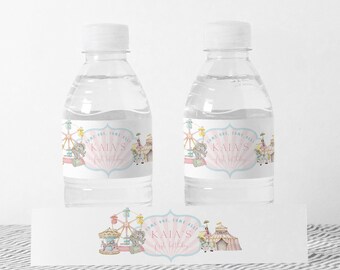 Puede incluir: Dos botellas de agua de plástico transparente con etiquetas rosas y azules que dicen "¡Venid todos! El 1er cumpleaños de Kaia". Las etiquetas presentan una ilustración caprichosa de una feria con una noria, un carrusel y un elefante. Una tercera etiqueta con el mismo diseño se muestra sobre una superficie blanca.