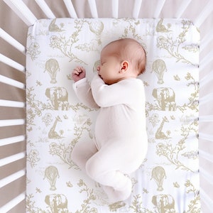 May include: A white crib sheet with a gold and white pattern of elephants, giraffes, hot air balloons, and flowers. A baby is sleeping on the sheet.
